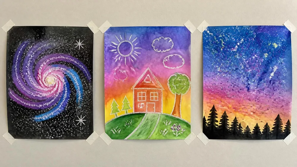 A row of finished kids' watercolor paintings taped to a wall: a salt galaxy, a crayon-resist scene, a splatter sky.