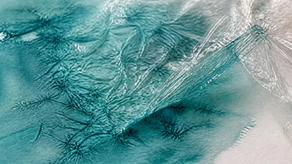 Crinkled plastic wrap pressed into a wet teal wash, one corner lifted to show the crystalline texture.