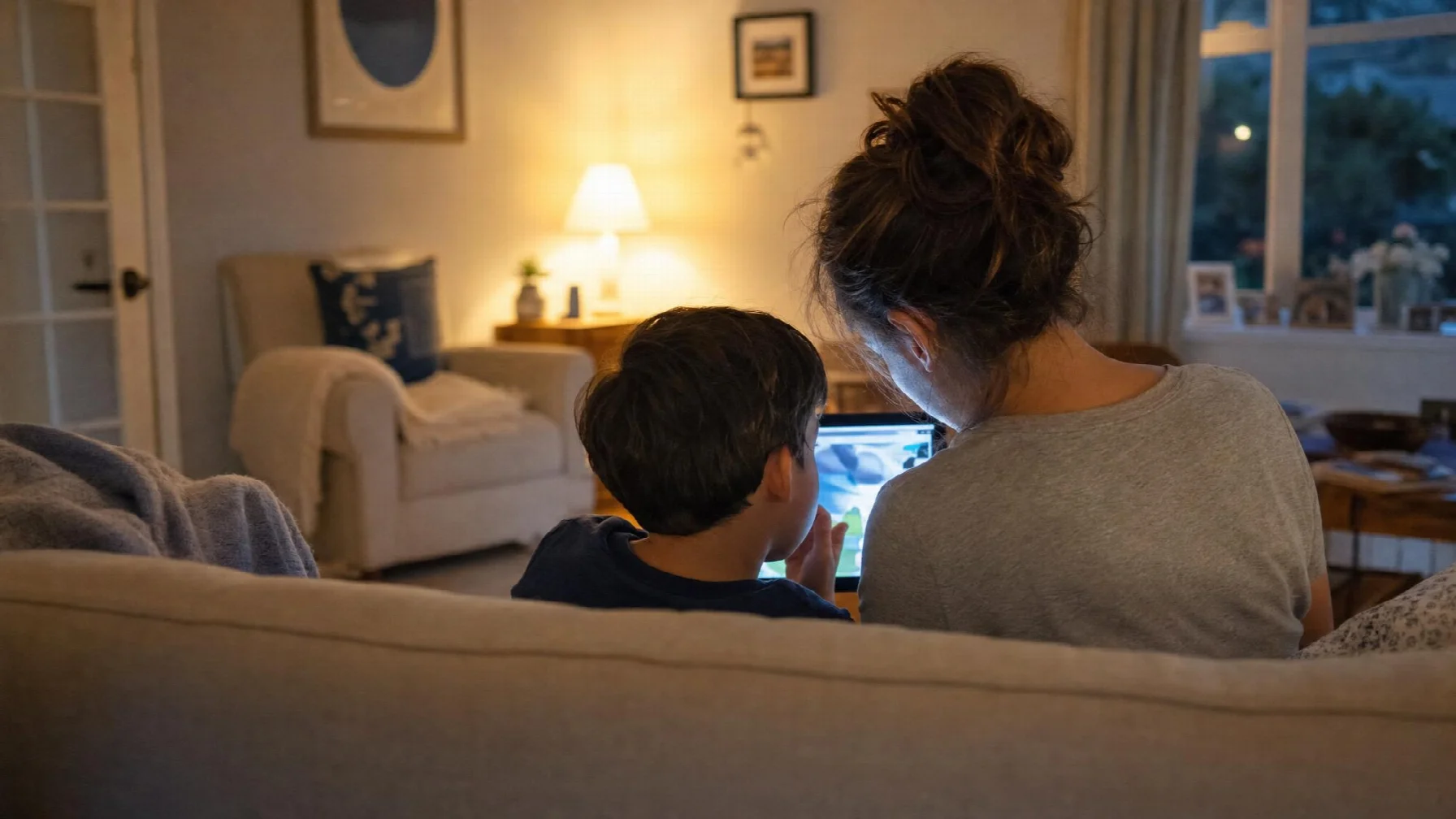 Parent and child on a couch in the evening, seen from behind, looking at a tablet together