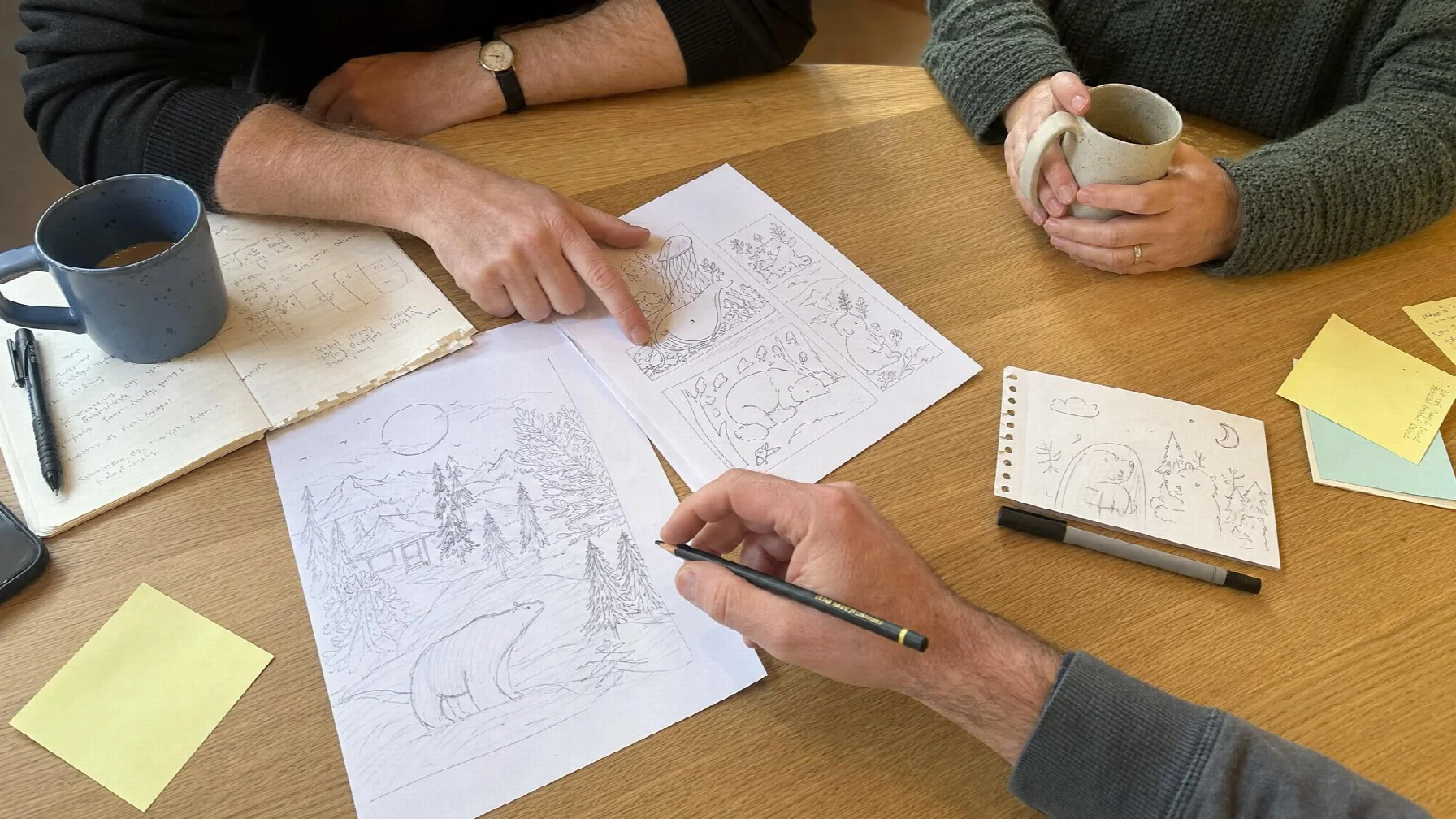 Adults' hands around a kitchen table covered with printed coloring pages, sketches, sticky notes, and coffee mugs