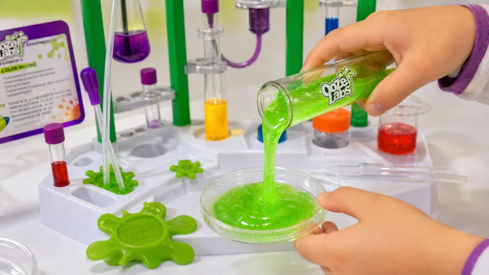 A child's hands tipping bright green glowing slime from an Ooze Labs test tube into a petri dish, the moulded lab station and pipettes in the background.