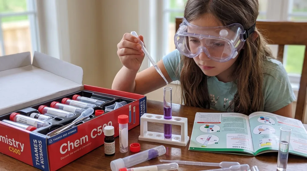 A ten-year-old in goggles adding drops from a pipette to a test tube of purple indicator solution, the open Thames and Kosmos Chem C500 kit and its manual on the table.