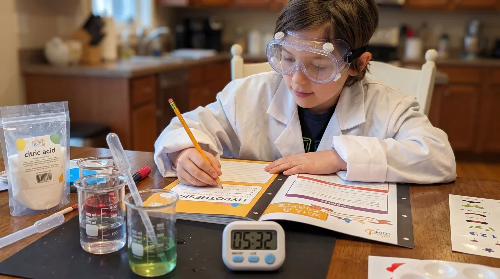 A nine-year-old in goggles writing a hypothesis in the Yellow Scope lab notebook, with beakers, a thermometer and a digital timer from the Beakers and Bubbles kit on the table.