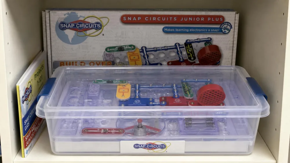 A Snap Circuits moulded storage tray sitting inside a clear lidded plastic tub on a shelf, with the original cardboard box flattened behind it and the project manual tucked down the side.