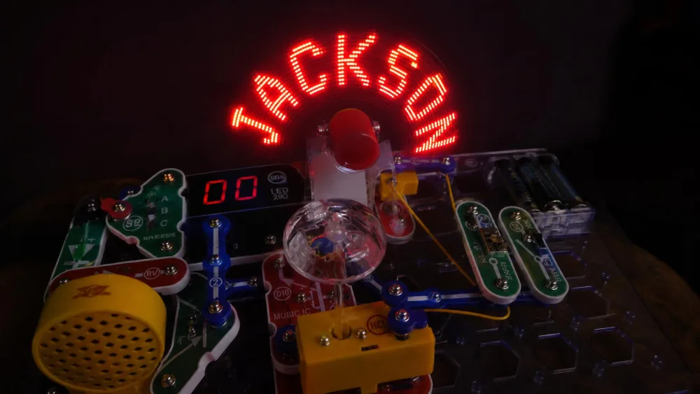A Snap Circuits Arcade SCA-200 spinning LED message fan photographed mid-spin in a dim room, showing a child's name written in light above the base grid.