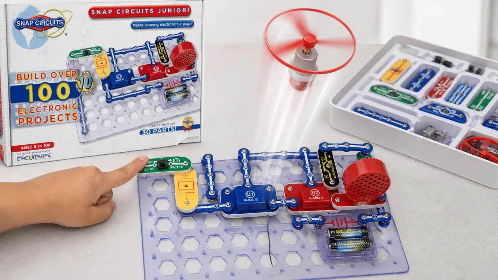 A Snap Circuits Jr. SC-100 flying-saucer project mid-launch above the blue base grid, with a child's finger still resting on the slide switch and the storage tray of parts beside it.