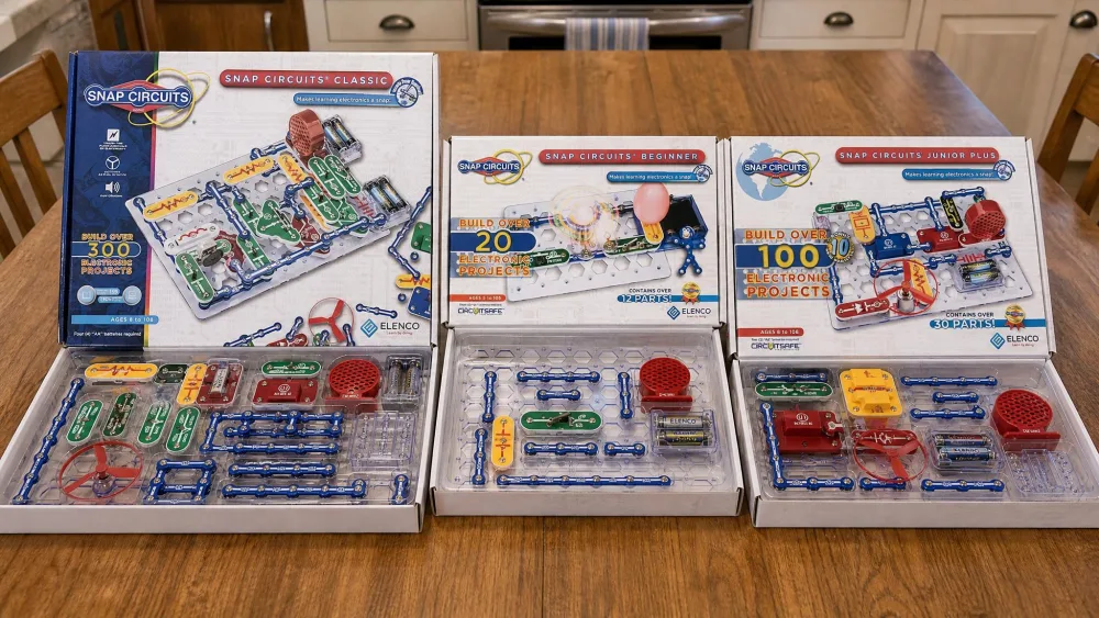 Three Snap Circuits boxes, the Classic SC-300, the Beginner SCB-20 and the Jr. SC-100, laid open side by side on a wooden kitchen table with their storage trays and colour-coded parts visible.