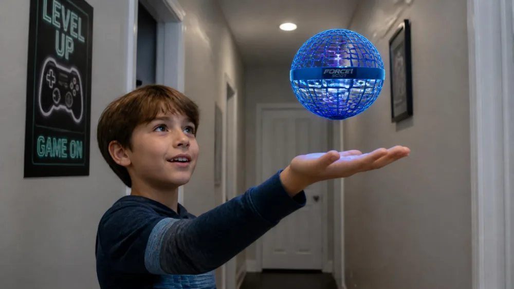 A child holding a flat palm under a glowing blue Force1 Scoot hand-operated drone, a round webbed plastic ball with LEDs lit inside, steering it upward in a hallway.