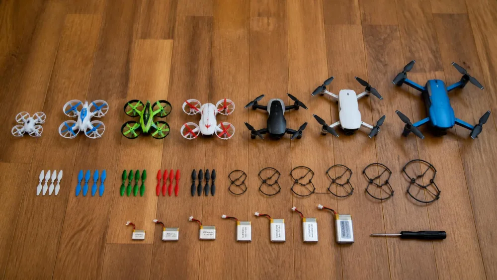 Seven kids' drones of different sizes in a row on a wooden floor next to spare propellers, guards, a screwdriver and several small batteries.