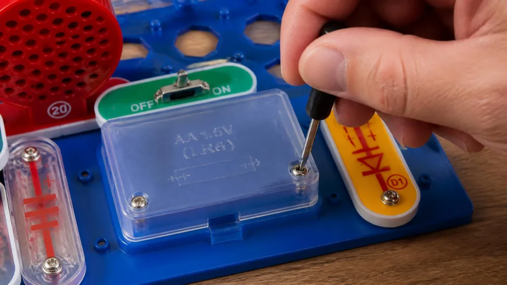 A close-up of an adult hand using a small screwdriver to open the screw-secured AA battery compartment on a children's electronics kit board.