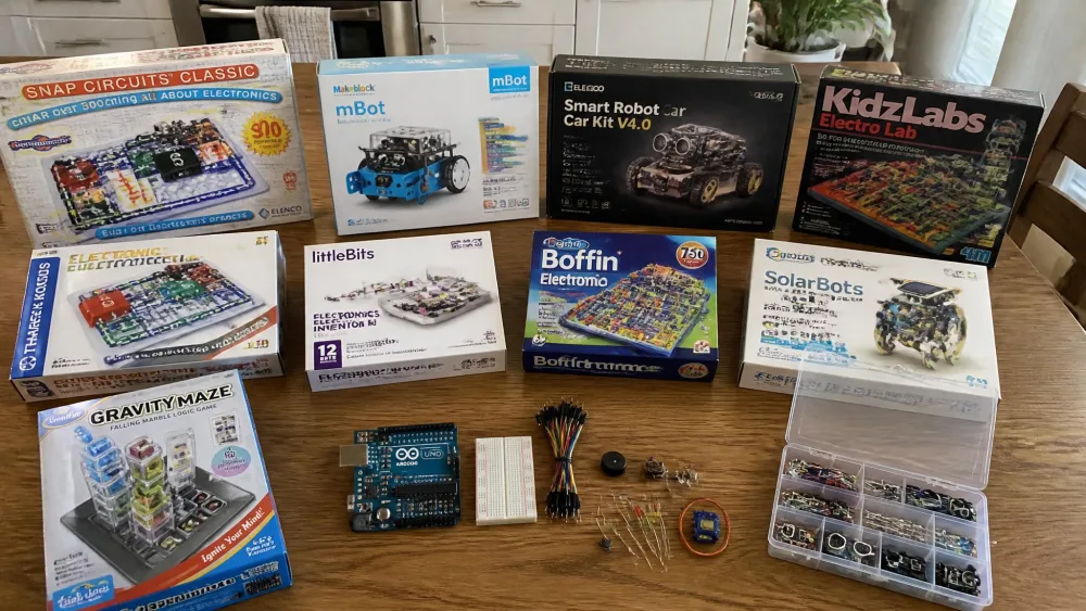 Ten children's electronics kits laid out across a wooden kitchen table, including Snap Circuits boxes, a Makeblock mBot robot, an ELEGOO robot car chassis and an Arduino board with loose components.