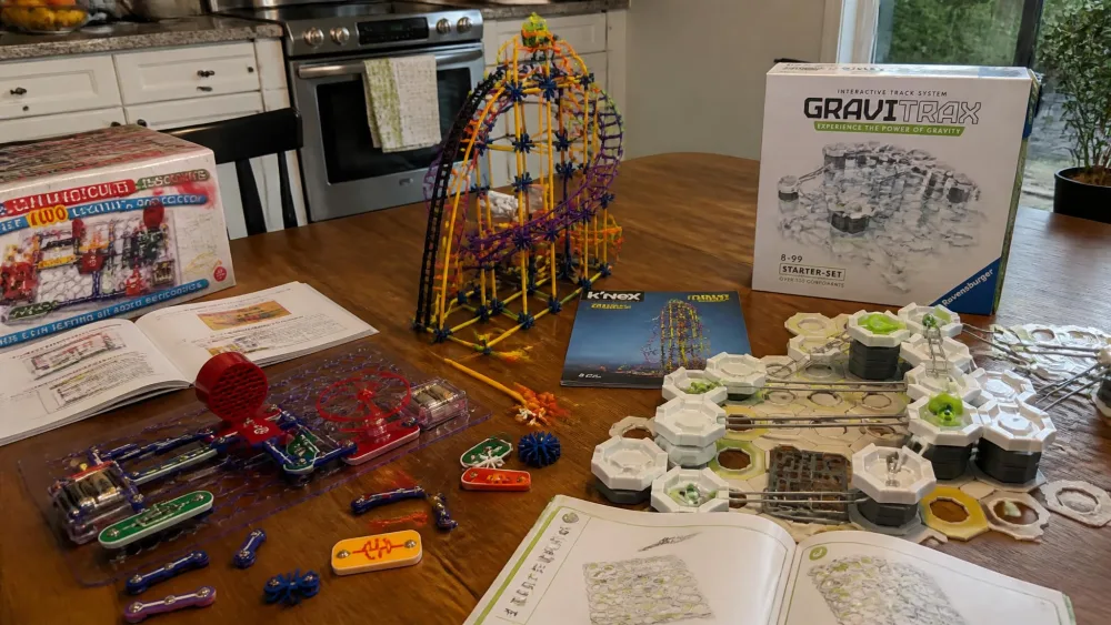 A kitchen table covered with partly built engineering kits, including a Snap Circuits baseplate, a half-built K'NEX coaster tower and a GraviTrax track.