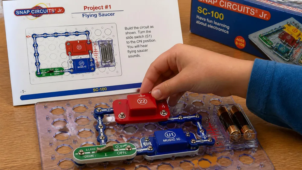 A child's hands snapping a red circuit block onto the grid baseplate of a Snap Circuits Jr. SC-100 kit, with the project manual propped up behind.