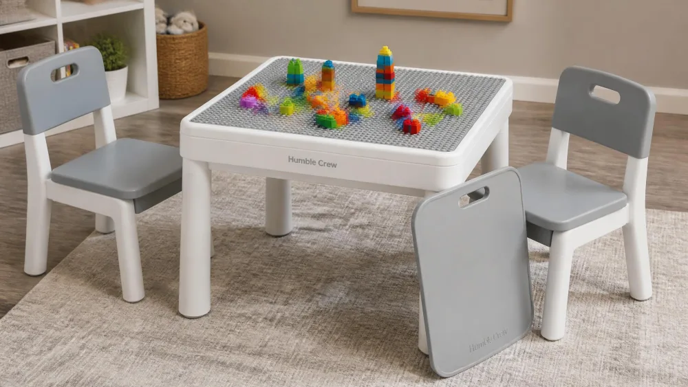 A small plastic Humble Crew activity table with two matching chairs, coloured blocks scattered on the studded surface and the smooth cover leaning against a leg.