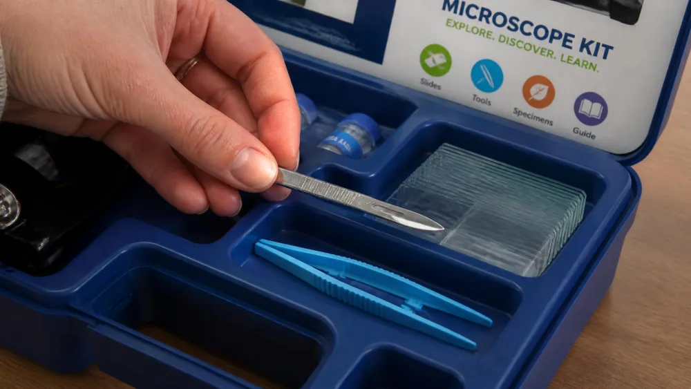The open tool tray of a kids' microscope kit showing a small metal scalpel, plastic tweezers and thin glass slides, a parent's hand lifting the scalpel.