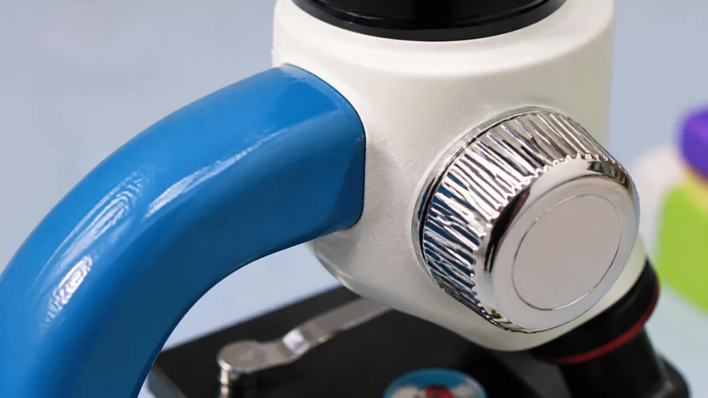 Close-up of a kids' compound microscope showing the join between a painted metal arm and a chrome-look plastic focus knob.