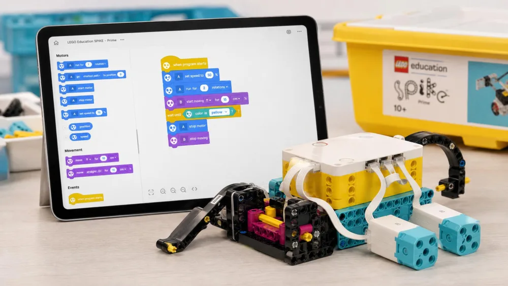 A partly assembled LEGO Education SPIKE Prime model with its yellow programmable Hub and two motors, a tablet behind it showing the block coding canvas.