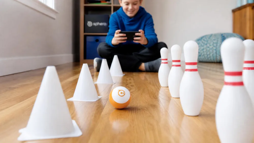 A Sphero Mini rolling between white traffic cones and bowling pins on a hardwood floor while a child steers it from a phone.