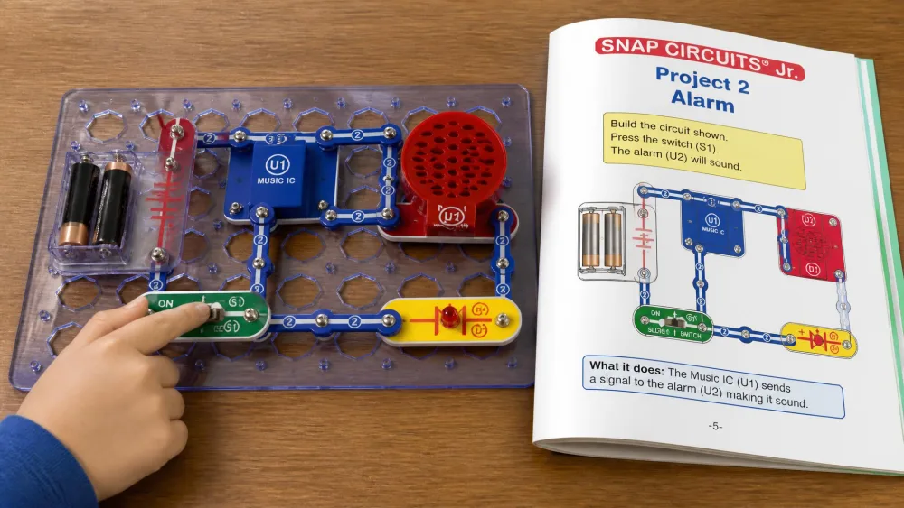 A completed Snap Circuits Jr. alarm circuit on its blue plastic grid, with a child's hand pressing the slide switch and the open project manual beside it.