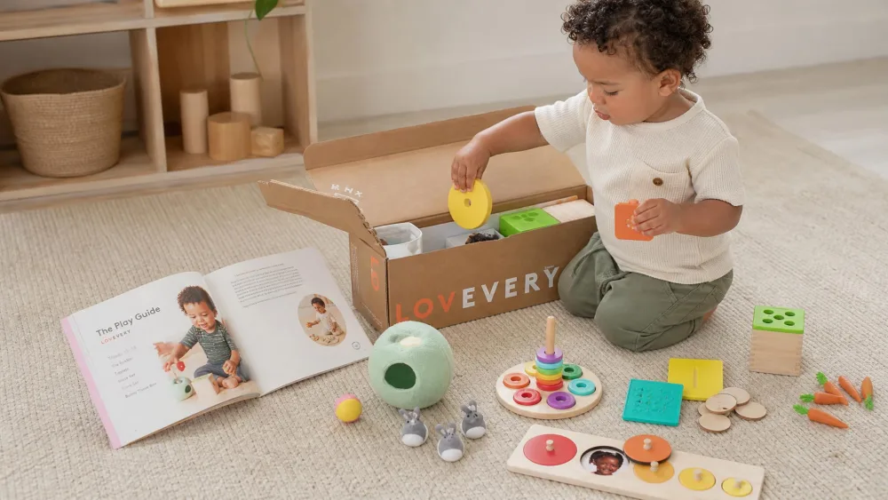 A toddler pulling wooden shapes out of an open Lovevery Play Kit box, with the Play Guide booklet open beside a fabric ball.