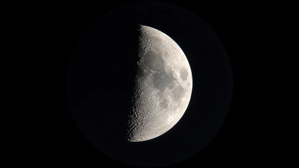 The view of a half moon through a beginner telescope eyepiece, craters casting long shadows along the terminator line between the lit and dark halves.