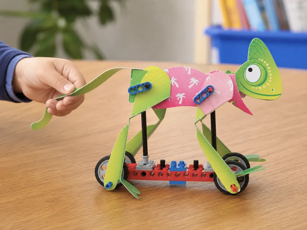 A child's hand pulling a finished Klutz LEGO Gear Bots lizard model across a wooden table.