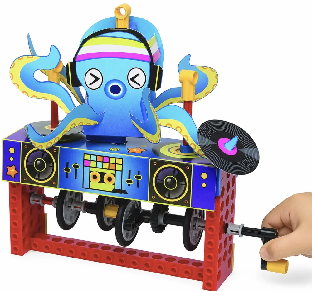 A child's hand turning the crank handle on a finished Klutz LEGO Gear Bots octopus model, the papercraft octopus skin spinning a small paper record on top of the LEGO Technic gear assembly.