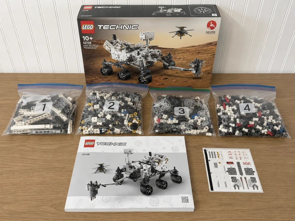 The LEGO Technic 42158 NASA Mars Rover Perseverance box contents laid out on a wooden table, showing the four numbered bags, the instruction booklet, and the small sticker sheet beside the closed box.