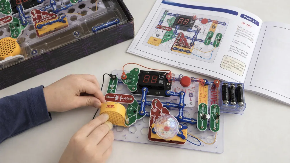 A child's hands pressing a yellow snap module onto the Snap Circuits Arcade grid board, with the open full-color manual showing a numbered circuit diagram beside it.