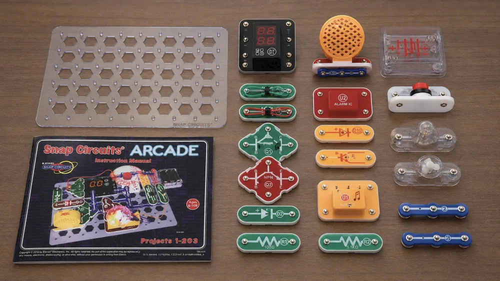 The Snap Circuits Arcade SCA-200 kit unpacked on a table, with color-coded snap modules, the clear grid base board, the dual-LED display, and the full-color manual.
