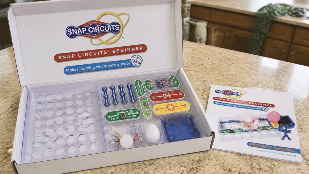 The open Snap Circuits Beginner SCB-20 box on a kitchen table, showing the base grid and color-coded snap parts beside the picture manual.