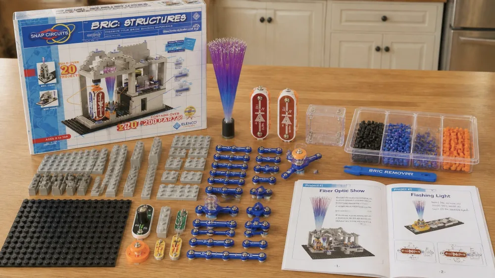 The contents of the Snap Circuits BRIC: Structures kit laid out on a wooden kitchen table, showing colored building bricks, blue Snap Circuits parts, Bric-2-Snap adapters, the brick-removal tool, and the open project manual.