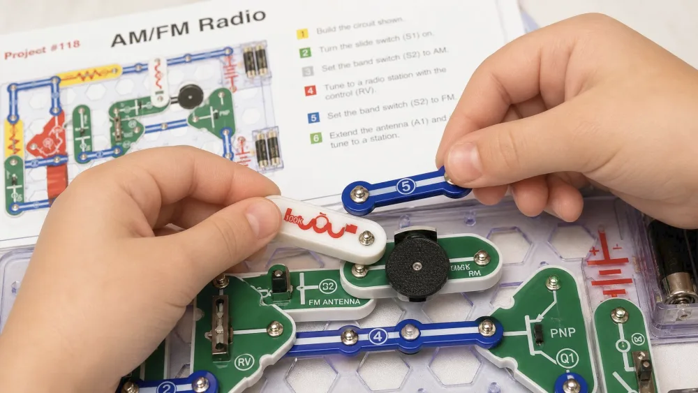 Close-up of a child's hands snapping an antenna part onto the Snap Circuits SC-300 grid while building the AM/FM radio project, with the numbered manual diagram behind.