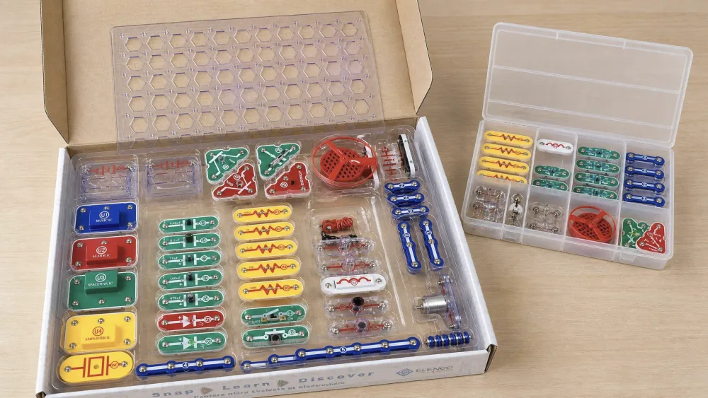 The Snap Circuits SC-300 storage tray open on a table, its 60-plus snap-together parts sorted into compartments, with a separate organizer box beside it.