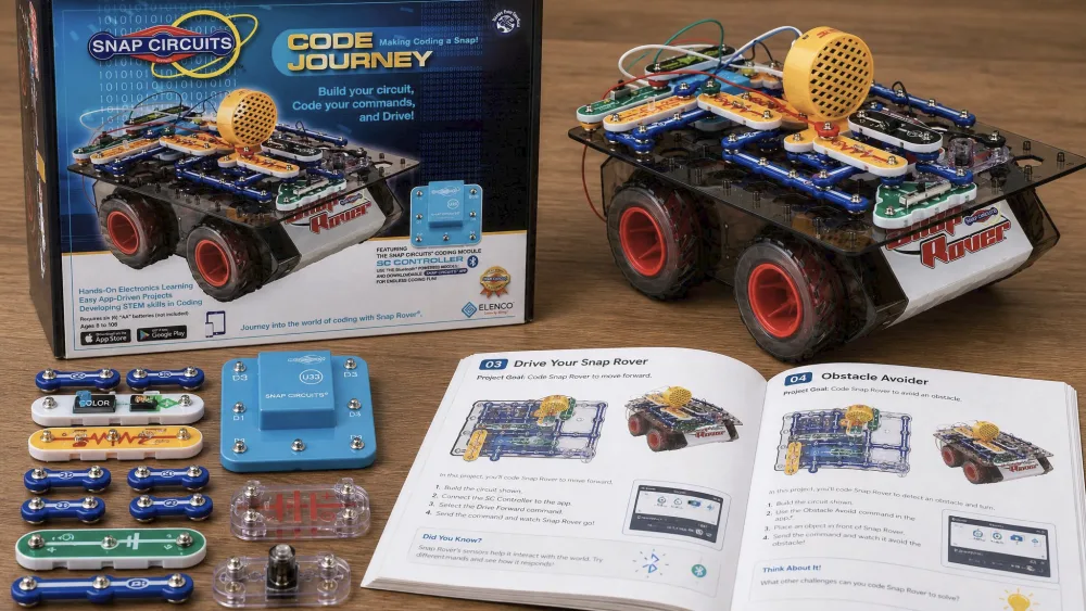 The Snap Circuits Code Journey kit laid out on a wooden table, showing the color-coded snap-together parts, the SC Controller module, and the built Snap Rover beside the open project manual.