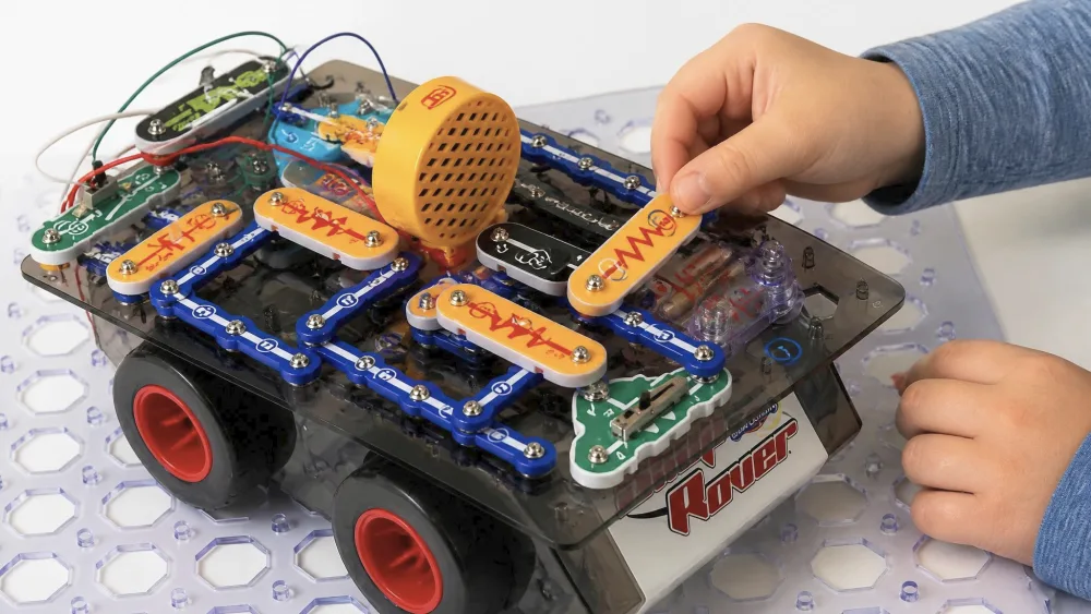 A child's hands placing the final color-coded part onto the assembled Snap Circuits Code Journey rover on a plastic build grid, with numbered parts clipped in place.