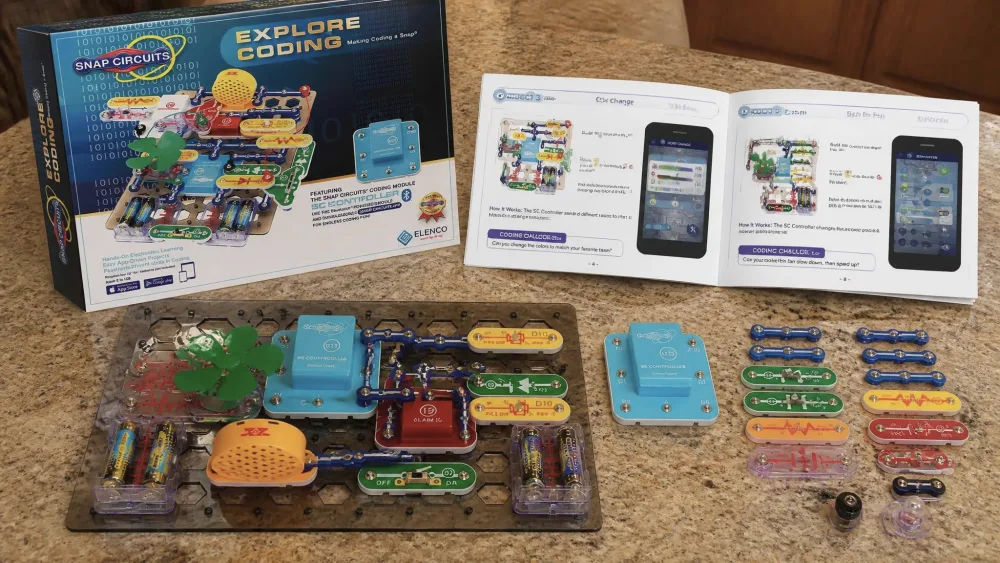 The Snap Circuits Explore Coding kit unpacked on a kitchen table, showing the base grid, color-coded snap parts, the SC Controller module, and the open project manual.