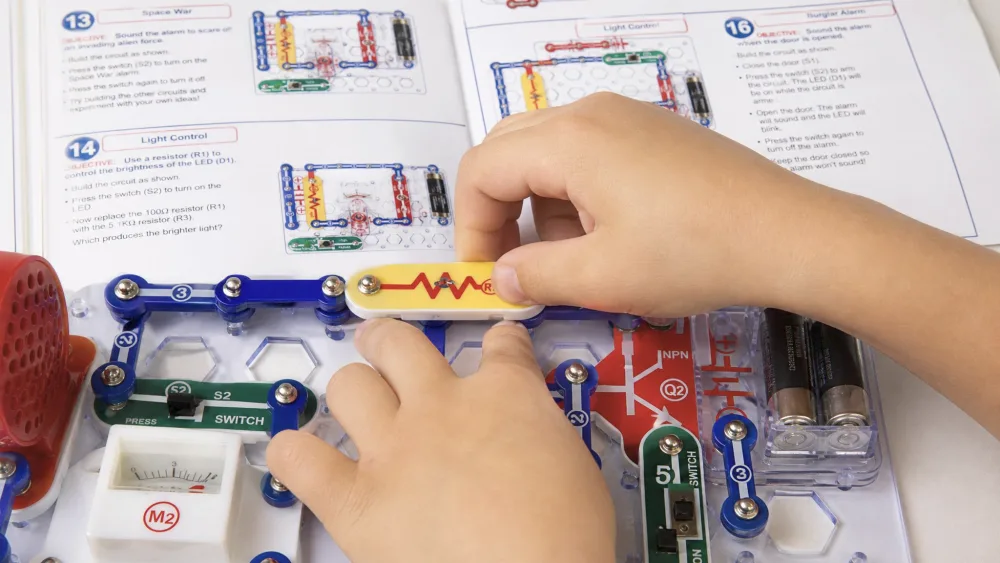 Close-up of a child's hands snapping a color-coded Snap Circuits part onto the white rows-and-columns base grid, with the numbered components and a full-color instruction manual visible in the background.
