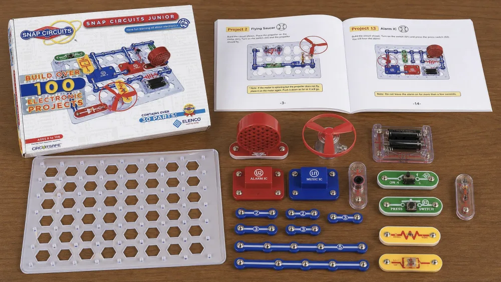 The contents of the Snap Circuits Jr. SC-100 on a wooden table: the white base grid, color-coded numbered snap parts, the motor with fan blade, speaker, switch, and the open full-color manual.