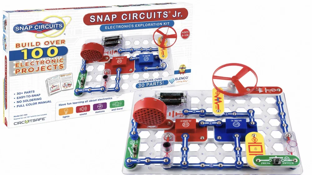Two children around age 8 leaning over a Snap Circuits Jr. SC-100 build on a kitchen table, one snapping a part onto the grid while the small motor fan spins.