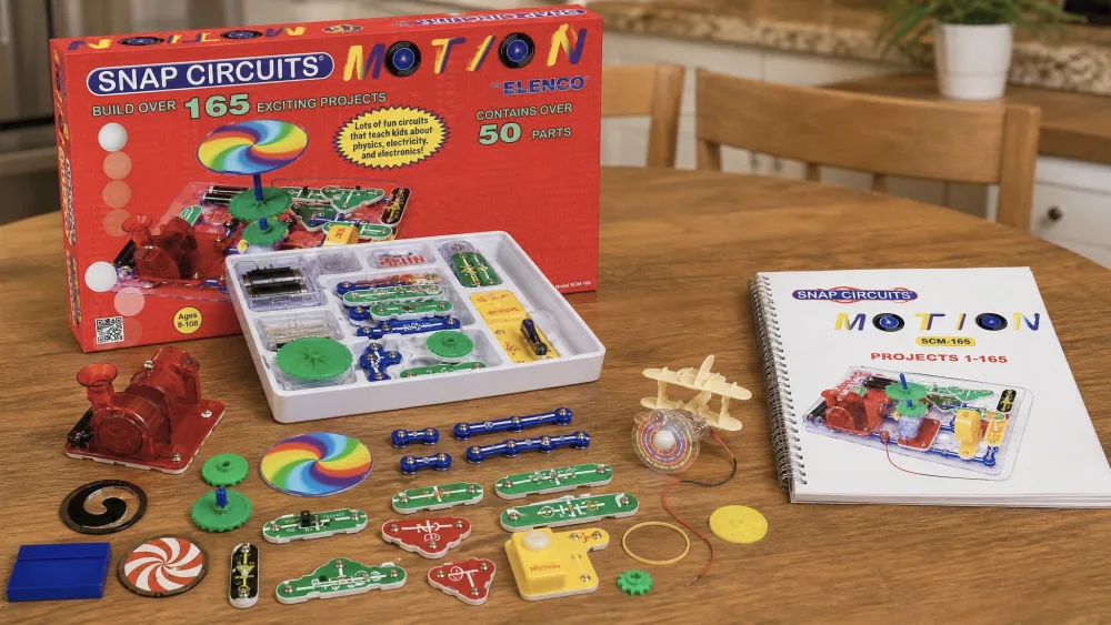 The open Snap Circuits Motion SCM-165 box on a wooden kitchen table with the color-coded snap parts, motor, gears, propeller, and spiral-bound project manual laid out beside it.