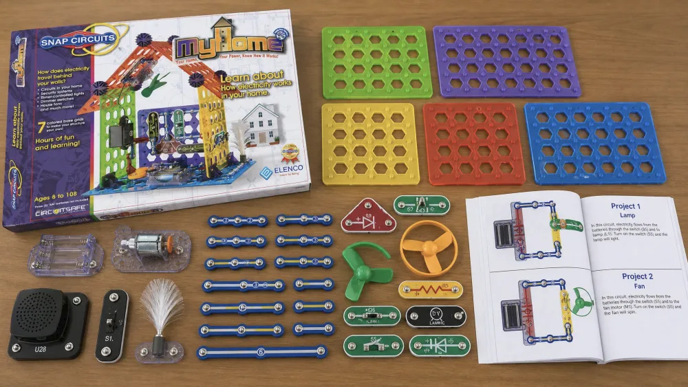 The contents of the Snap Circuits MyHome SC-MYH7 kit laid out on a table, showing colored base grids, snap blocks, wires, a sound module, a motor, and the open manual.
