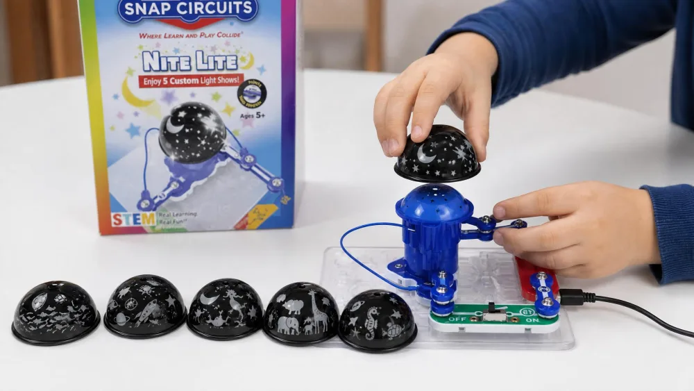 A child's hands lifting one themed projection dome off the assembled Snap Circuits Nite Lite to swap in another, with the five domes lined up on the table beside the USB-powered unit.