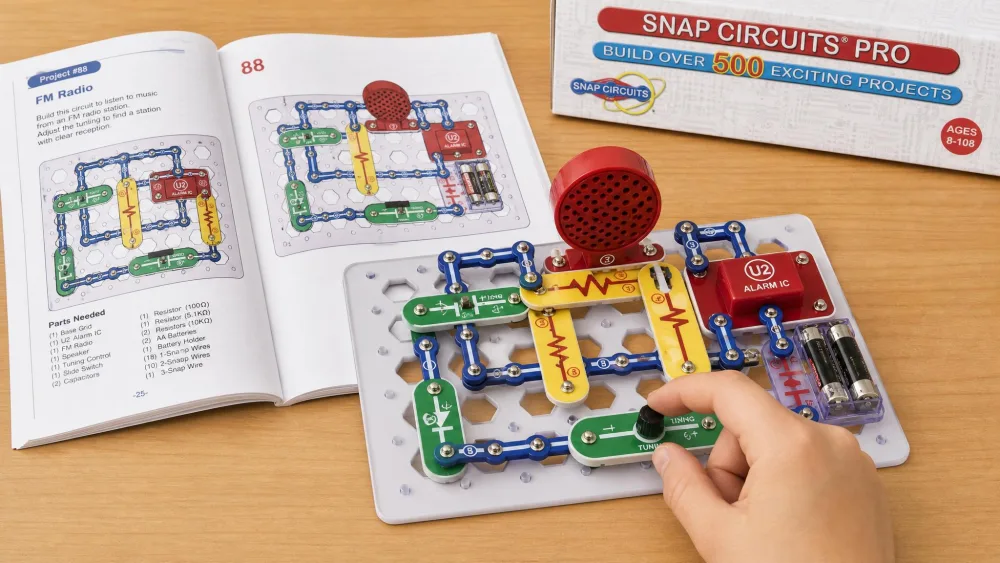 A completed Snap Circuits Pro FM radio circuit on the white grid base, with a child's hand adjusting the tuning while the open manual shows the matching numbered build diagram.