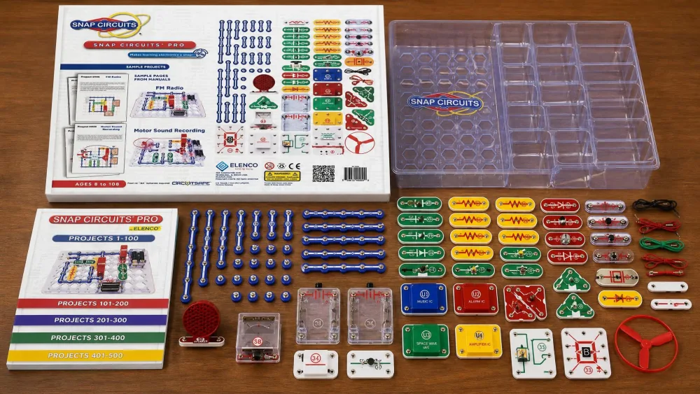 The full set of Snap Circuits Pro SC-500 components laid out on a table beside the included storage tray, showing the color-coded numbered snap modules, the wires, and the stack of full-color project manuals.