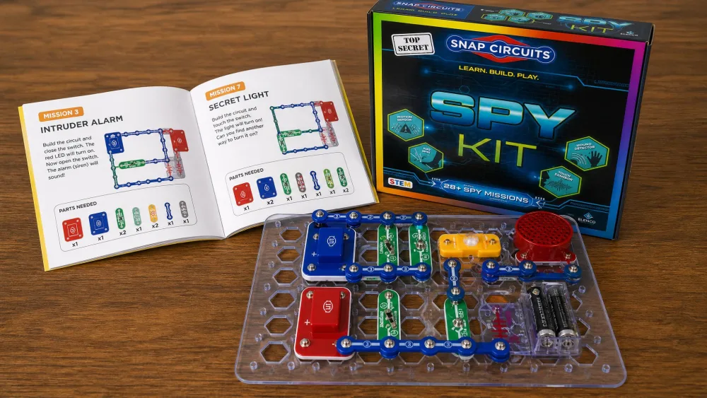 The Snap Circuits Spy Kit base grid with color-coded snap-together circuit parts and the open full-color picture manual laid out on a wooden table, ready to build a spy gadget.