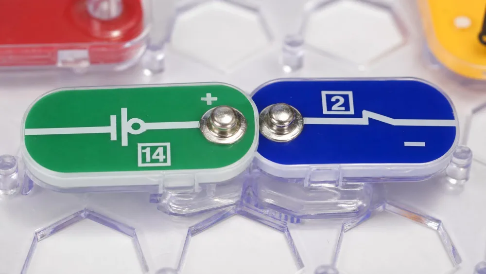 Close-up of two color-coded Snap Circuits Spy Kit parts snapping together at their metal contacts on the plastic grid, showing the no-solder connection.