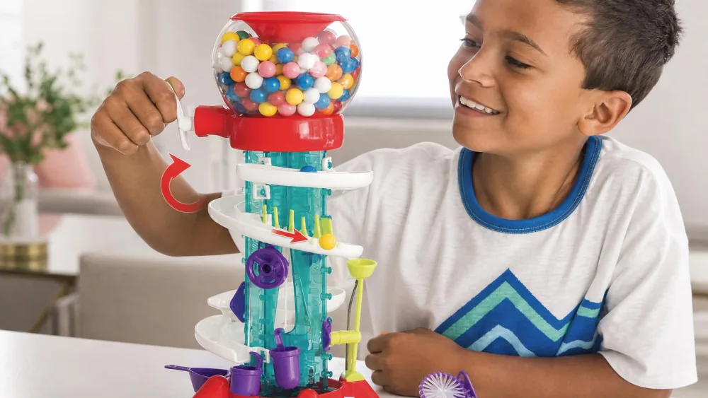 A child's hand turning the hand-crank on a built Thames & Kosmos Gumball Machine Maker, dispensing a yellow gumball into a track segment.