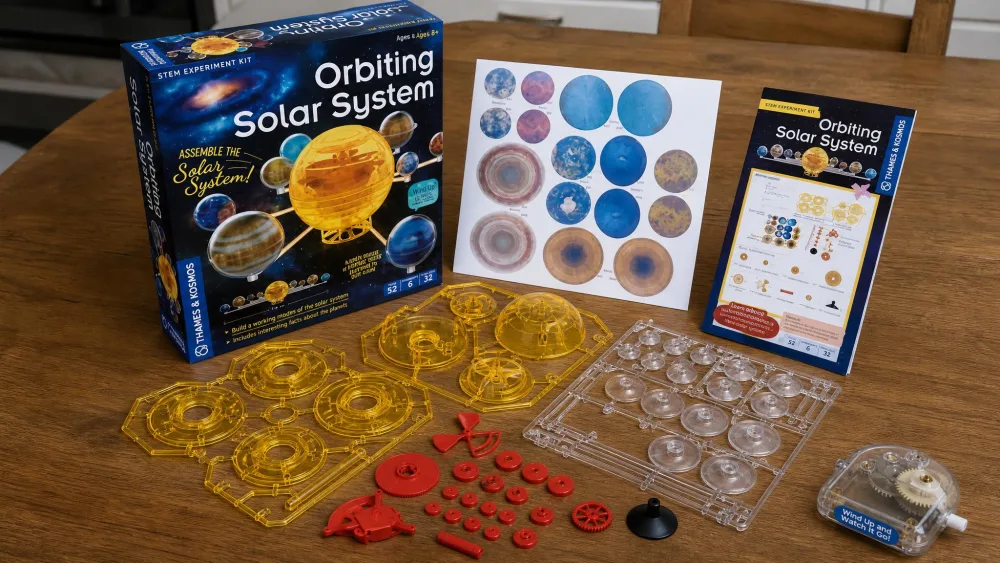 The unboxed Thames & Kosmos Orbiting Solar System kit laid out on a wooden kitchen table, showing the windup motor unit, plastic part frames, printed planet graphics, and the full-color instruction booklet.