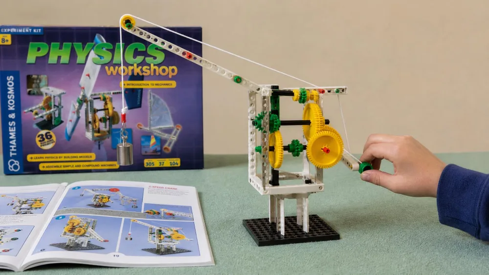 A completed two-speed crane built from the Thames & Kosmos Physics Workshop kit, with a child's hand turning the gear crank to lift a small weight on the pulley line.